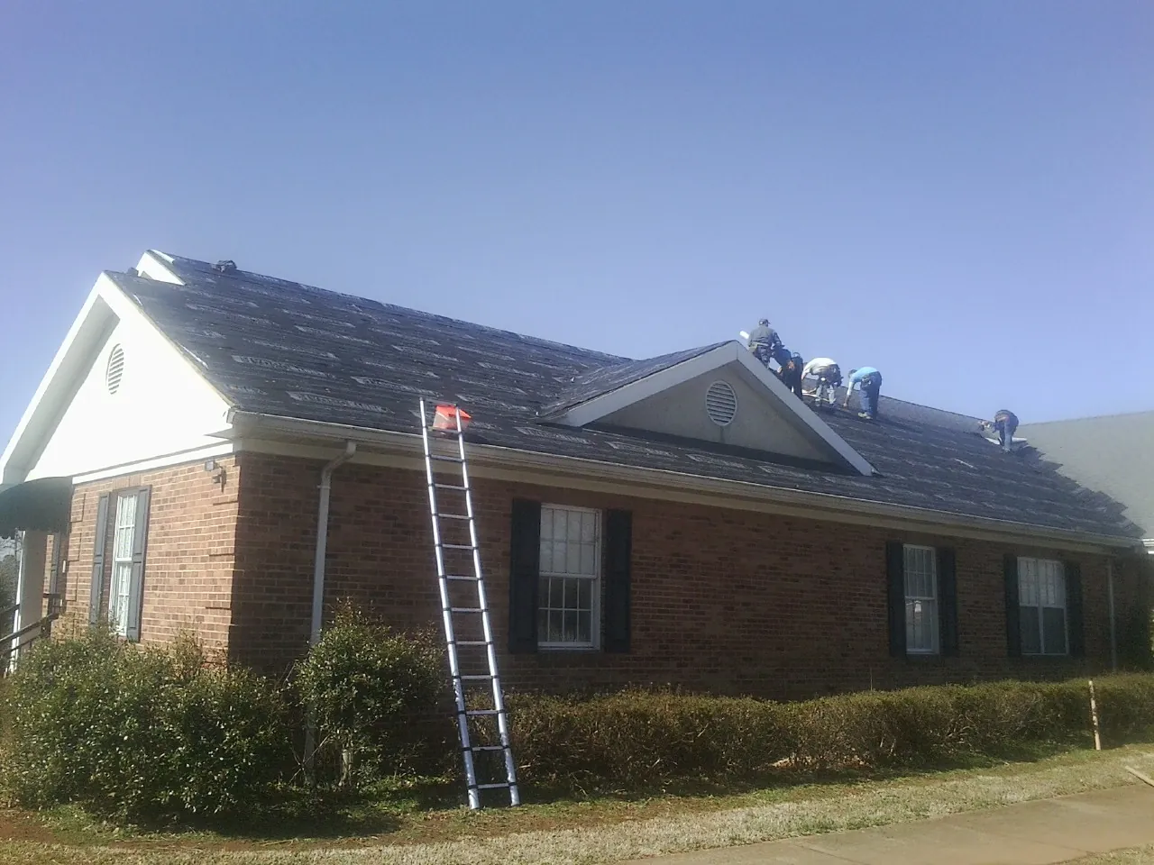 Crew tearing shingles off a brick ranch roof with a ladder set — Emergency Roof Repair work by our crew in Chapin