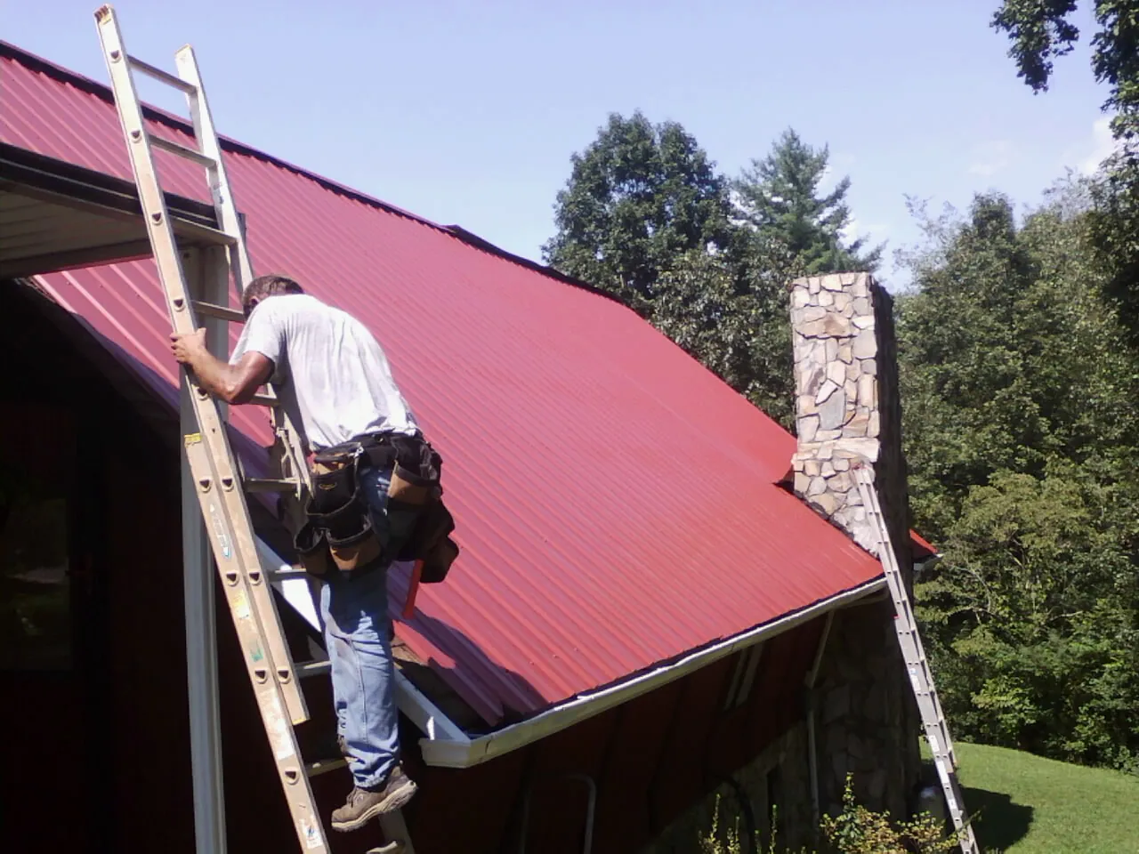 Roofer on a ladder beside a red metal roof and stone chimney — Soffit & Fascia Repair by Roofing Brothers in Chapin